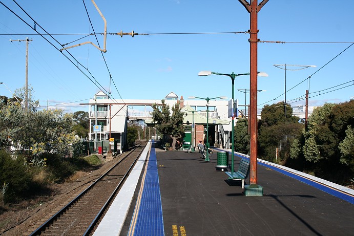 The view looking along the platform after its upgrade to an 'Easy Access Station' during 2005. The platform has been resurfaced and all gardens removed except for the old Banksia tree near the western end of the building. Tactile tiles have been installed on the platform edges, the waiting room floor was lowered to enable wheelchair access as well as an easy access toilet installed. Also prominent are the tops of the lift towers to the platforms and to Wilson Way (up side) and the covered walkways and stairs.