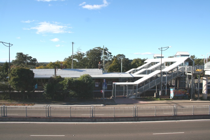A photo of Blaxland Station taken from the footbridge over the Great Western Highway near the shops. It is looking at Platfrom 2 and the Down Main, and shows the new covered walkways and stairs, as well as the tops of the lift towers to the platforms and Wilsons Way on the far side. Access to the shops remains by way of a ramp leading from the bridge over the highway.