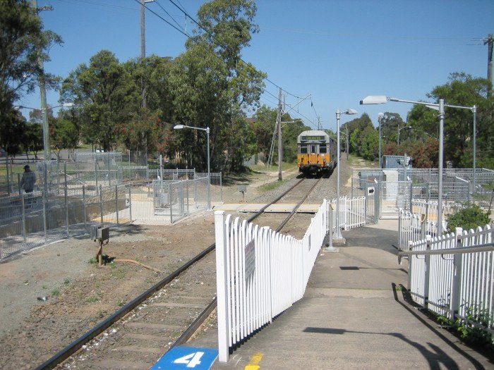A four car suburban set approaches Telopea station on its way to Carlingford. The newly installed pedestrian crossing, with train activated automatic gates and emergency exits, are shown at the end of the platform.
